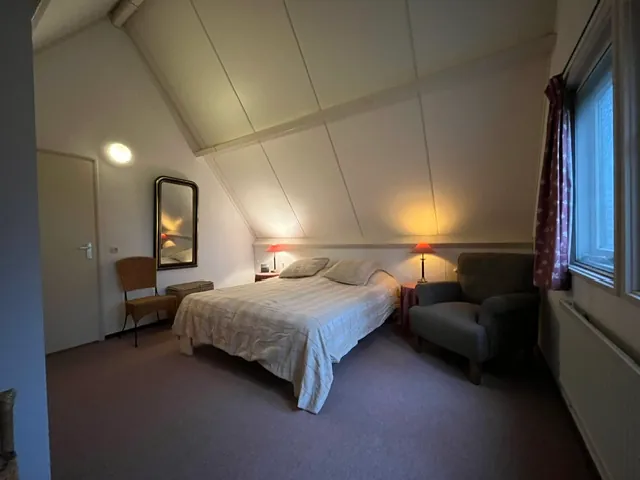 Een andere slaapkamer onder het schuine dak: een breed bed tussen twee lampjes en een leunstoel bij het raam.